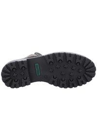 Suela de zapato de goma negra con un patrón de tracción profundo, que presenta una etiqueta de marca en verde y negro. La superficie muestra un acabado texturizado y duradero.