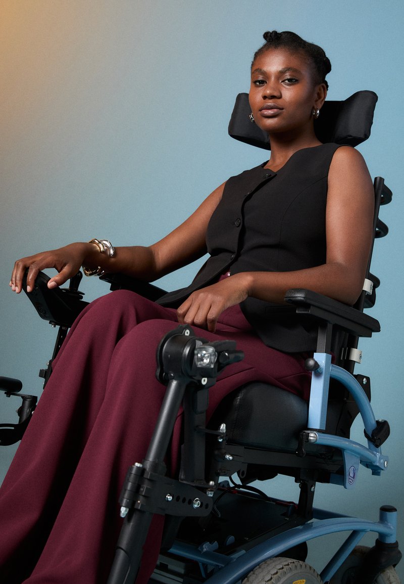 Fauteuil roulant électrique avec un siège noir et des composants réglables, doté d'un cadre bleu clair et de pantalons évasés bordeaux. Utilisateur avec des cheveux tressés.