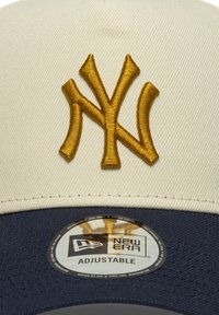 Cremefarbene Baseballkappe mit dunkelblauem Schirm. Verfügt über ein gesticktes goldfarbenes NY-Logo. Das Innenetikett enthält "New Era" und "Verstellbar".