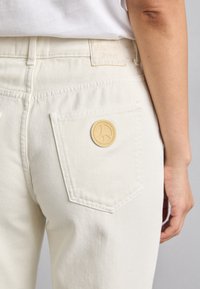 Baltie džinsi ar aizmugurējo kabatu, kurai ir apaļa brūna uzlīme, kurā iegravēts miera simbols. Minimālisma dizains un šuvju detaļas.