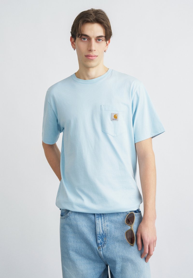 Νέος άνδρας που φοράει ανοιχτό μπλε T-shirt Carhartt και τζιν, με γυαλιά ηλίου στερεωμένα στην τσέπη, στέκεται μπροστά σε απλό λευκό φόντο.