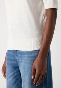 Bras droit d'une personne portant un pull blanc à côtes et un jean en denim bleu sur un fond neutre.