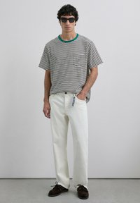 Camiseta de rayas con cuello verde, jeans blancos y zapatos de ante marrón. El modelo lleva gafas de sol negras; ajuste relajado y bolsillo en la camiseta.