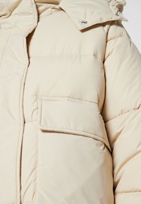 Veste d'hiver beige matelassée avec grande poche frontale et bouton-pression sur le col, montrant les détails des coutures et la texture du tissu.