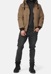 Braune gesteppte Jacke mit Kapuze, schwarzer Pullover darunter, graue Jeans und schwarze Schnürstiefel. Verfügt über Reißverschlusstaschen und Toggle-Akzente.