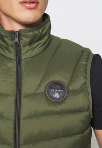 Olijfgroene puffer vest met hoge kraag en rits, voorzien van een ronde zwarte patch met oranje en witte details op de borst.