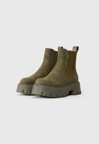 Un paio di stivaletti Chelsea in suede verde oliva con suole a piattaforma spesse e pannelli laterali elasticizzati a coste su uno sfondo chiaro.