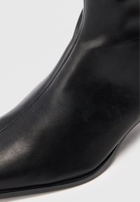 Botte de cheville en cuir noir avec un bout pointu et une texture lisse sur un fond clair.