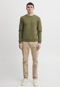 Hanorac verde măsliniu cu decolteu rotund, textură moale și detaliu cu logo alb. Asortat cu pantaloni bej și adidași albi.