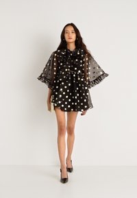 Μαύρο φόρεμα με πουά, με κολάρο με φουσκωτό σχέδιο και διαφανείς μανίκες σε σχήμα καμπάνας, διαθέτει φουσκωτό φούστα και υφή. Φορεμένο με μαύρες γόβες.