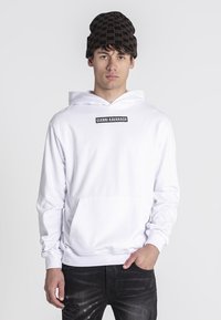 Biele mikina s kapucňou a predným vreckom, s čiernym logom na hrudi. Doladené s tmavými roztrhnutými džínami a vzorovanou hnedou čiapkou.