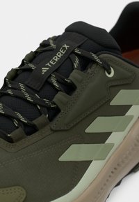 Πράσινο παπούτσι πεζοπορίας με υφή υφάσματος στο πάνω μέρος, σκούρες λεπτομέρειες, ενισχυμένη μύτη και διακριτικό γκρι λογότυπο και ρίγες στο πλάι.