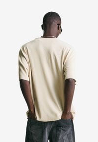 Bež majica s kratkimi rokavi in okroglim izrezom, narejena iz teksturiranega materiala. Nošena z temno sivohlačami, roke v žepih.