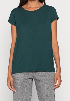 Frau trägt ein dunkelgrünes T-Shirt mit kurzen Ärmeln und hellgraue Jeans, mit einem Line-Art-Tattoo am linken Unterarm.