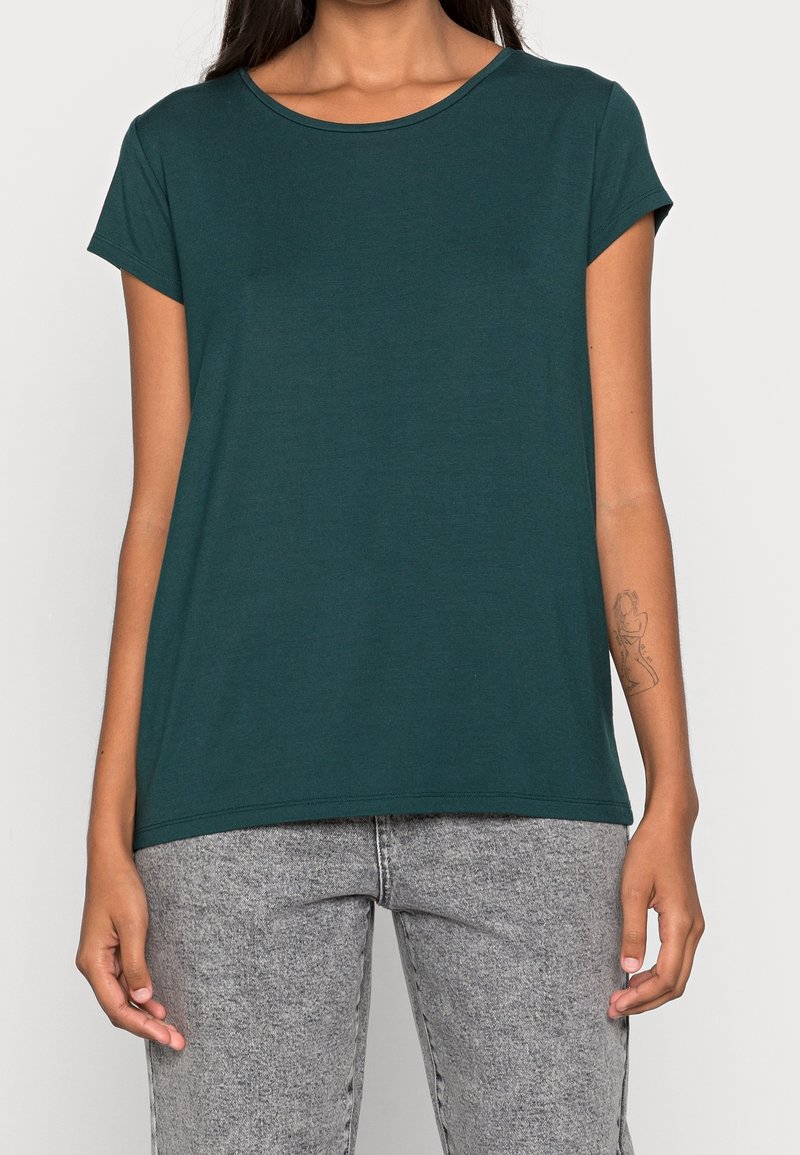 Femme portant un t-shirt vert foncé à manches courtes et un jean gris clair, avec un tatouage en ligne artistique sur son avant-bras gauche.
