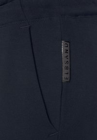 Marineblaue Sweatpants aus weicher Baumwolle mit glatter Textur. Enthält ein kleines schwarzes Stoffetikett, das an der Seite angenäht ist.