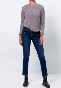 Mujer vistiendo una blusa de cuadros púrpura y blanca de manga larga, jeans ajustados azul oscuro y botas negras hasta el tobillo, de pie contra un fondo blanco.