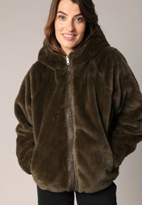 Veste zippée en fausse fourrure vert olive foncé, dotée d'une capuche, avec une texture pelucheuse et une coupe décontractée. Manches longues et poches avant visibles.