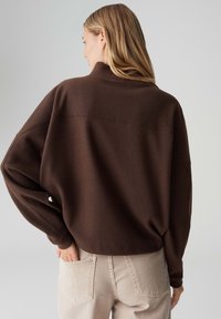Bruine oversized trui met een hoge kraag en verlaagde schouders, textuurstof, gedragen met lichtbeige broek, achteraanzicht.