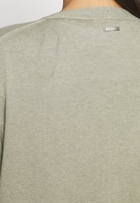 Pull en maille vert clair avec un col rond et un petit logo en métal au dos. Texture lisse avec un léger motif en maille.