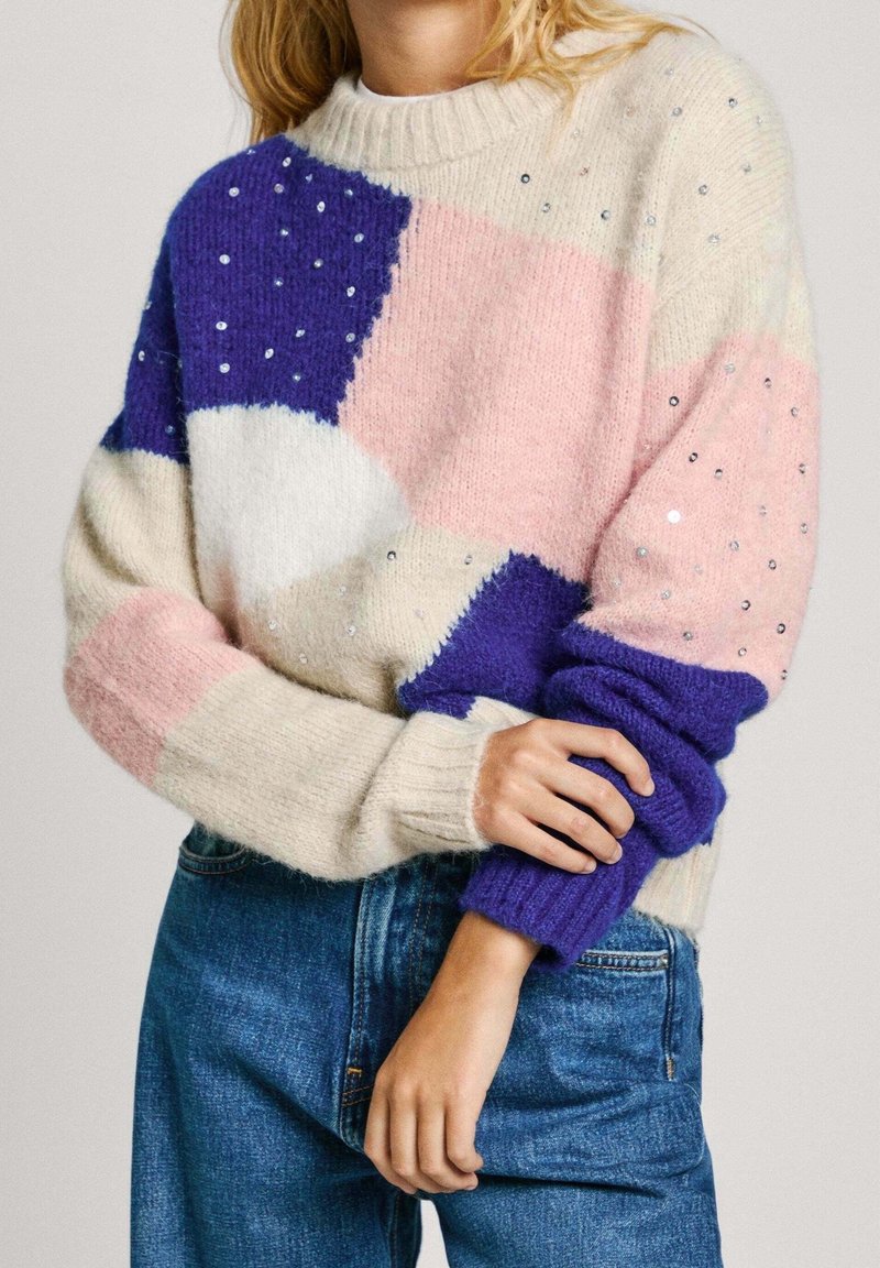 Femme portant un pull multicolore aux sections violettes, roses, blanches et crème ornées de petits sequins brillants, associé à un jean bleu.