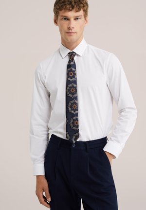 Hombre con camisa blanca, corbata azul marino con patrón y pantalones oscuros, de pie con una mano en el bolsillo delante de un fondo liso.