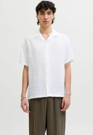 Junger Mann mit kurzem lockigem Haar, trägt ein weißes kurzärmeliges Hemd und lockere braune Hose, steht vor einem schlichten Hintergrund.