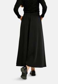 Melnas, plūstošas maksi svārki ar elastīgu jostu, izgatavoti no gludas auduma. Raksturīga vaļīga piegriezuma un minimālistiska dizaina. Redzamas melnas kurpes.