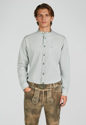 Junger Mann trägt ein hellgraues Hemd mit Knöpfen und traditionelle braune Lederhose mit Stickerei sowie eine dekorative Gürtelschnalle.