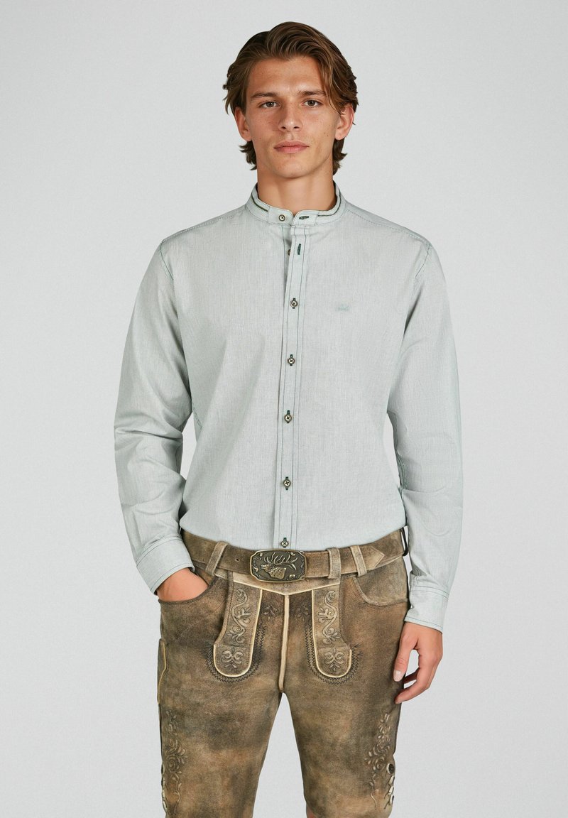 Junger Mann trägt ein hellgraues Hemd mit Knöpfen und traditionelle braune Lederhose mit Stickerei sowie eine dekorative Gürtelschnalle.