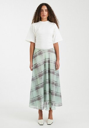 Vrouw met krullend haar draagt een wit topje met korte mouwen en een lange lichtgroene geruite rok met witte puntige schoenen, staand tegen een witte achtergrond.