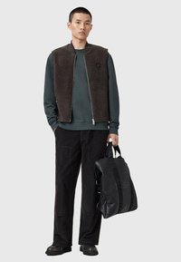 Hnědá fleecová vesta přes tmavozelenou mikinu, v kombinaci s černými kordovým kalhotami a černými koženými botami. Drží strukturovanou černou tašku.