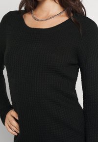 Haut à manches longues texturé noir avec un col en arrondi. Présente un motif côtelé et une coupe ajustée. Un collier chaîne argentée épaisse est porté.