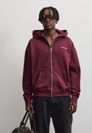 Uomo con capelli intrecciati che indossa grandi occhiali da sole, una felpa con cappuccio bordeaux con zip, jeans scuri, collane d'oro e tiene una borsa in pelle marrone.