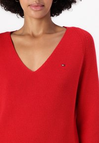 Maglione rosso lavorato a maglia con scollo a V e trama a coste. Presenta un piccolo logo sul petto sinistro superiore. Maniche lunghe.