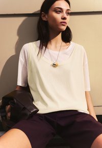 T-shirt bianco largo indossato sotto una canotta gialla chiara, abbinato a pantaloni corti viola scuro e una borsa in pelle marrone. Collana in argento e giallo.