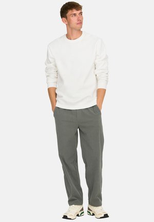 Man wearing a white long-sleeve sweatshirt, gray pants, and beige sneakers, standing hands in pockets, looking to the right.