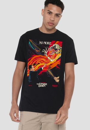 Junger Mann trägt ein schwarzes T-Shirt mit einem Stranger Things Upside Down Kapitel acht Motiv, das einen Jungen zeigt, der Kräfte gegen ein Monster einsetzt, mit dem Text "No More."