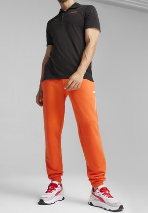 Svart pikétröja med röd märkning, kombinerad med klargula sweatpants och vita sneakers med röda detaljer. Enkel, atletisk design.