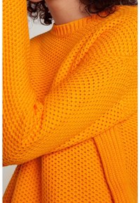 Donna che indossa un maglione lavorato a maglia di colore arancione acceso con maniche lunghe, mostrata dal collo alla vita con un braccio alzato.