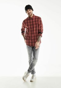 Červená a černá kostkovaná košile, šedé distressed džíny, bílé tenisky a černá pletená čepice. Viditelné tetování na pažích. Pozice vestoje.