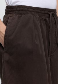 Gros plan sur un pantalon à taille élastique marron foncé avec des poches latérales et une chemise blanche rentrée. La main repose dans la poche.