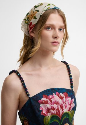 Νεαρή γυναίκα με ξανθά μαλλιά φοράει φλοράλ μαντήλι στο κεφάλι και μπλε φόρεμα με ένα μεγάλο ροζ λουλούδι και μαύρα ιμάντες με χάντρες.