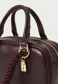 Dunkelbraune Lederhandtasche mit zwei Griffen, goldfarbenem Reißverschluss und goldfarbenem Vorhängeschloss, das an einem geflochtenen burgunderfarbenen Riemen befestigt ist.