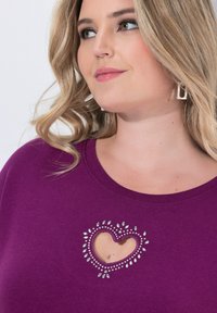 Donna con capelli biondi mossi, che indossa un top viola con un taglio a forma di cuore adornato con piccole borchie trasparenti e orecchini geometrici in argento.