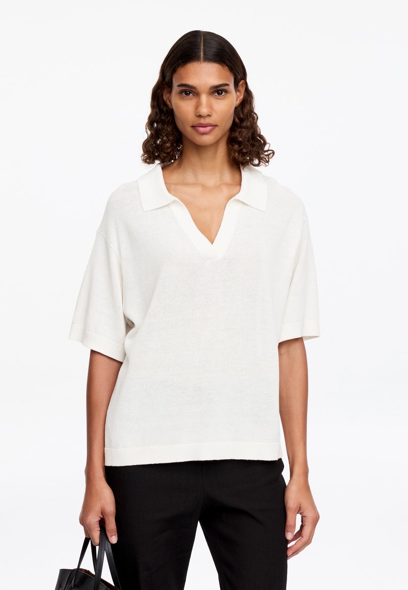 Femme aux cheveux bouclés porte un polo en maille blanc ample à manches courtes et un pantalon noir, tenant un sac à main noir à ses côtés.