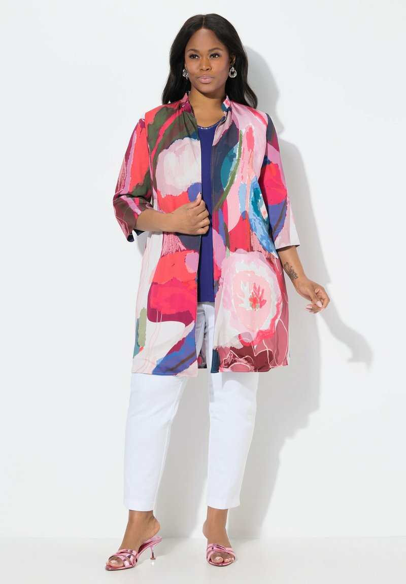 Kolorowa kurtka w stylu kimono z kwiatowymi wzorami w odcieniach różu, niebieskiego i zielonego, noszona na granatowej bluzce, w połączeniu z białymi spodniami i różowymi sandałami.