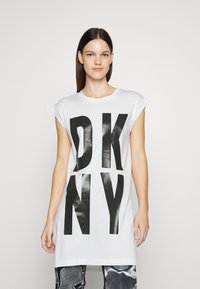 Balta medvilnė marškinėliai su dideliu juodu "DKNY" spaudiniu. Trumpi rankovės, laisvas siluetas, tiesus apatinis kraštas. Fonas paprastas ir šviesus.