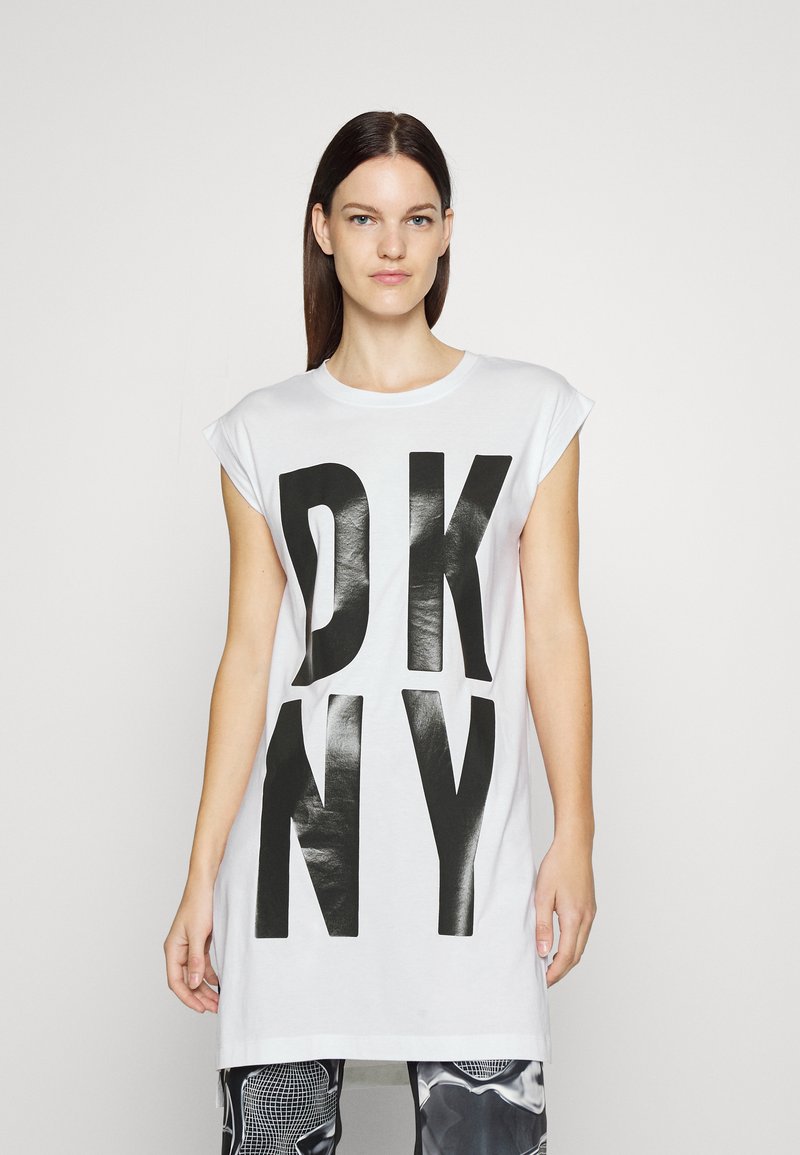 Balta medvilnė marškinėliai su dideliu juodu "DKNY" spaudiniu. Trumpi rankovės, laisvas siluetas, tiesus apatinis kraštas. Fonas paprastas ir šviesus.