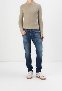 Beżowy sweter w prążki i wyblakłe niebieskie dżinsy. Sneakersy są białe z czarnymi paskami i brązowymi podeszwami. Model stoi z jedną ręką w kieszeni.